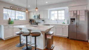 A modern kitchen with white cabinets, a large island with stools, a stainless steel refrigerator, and a sign that reads "BEACH HOUSE". - Pacific Pearl (San Diego)