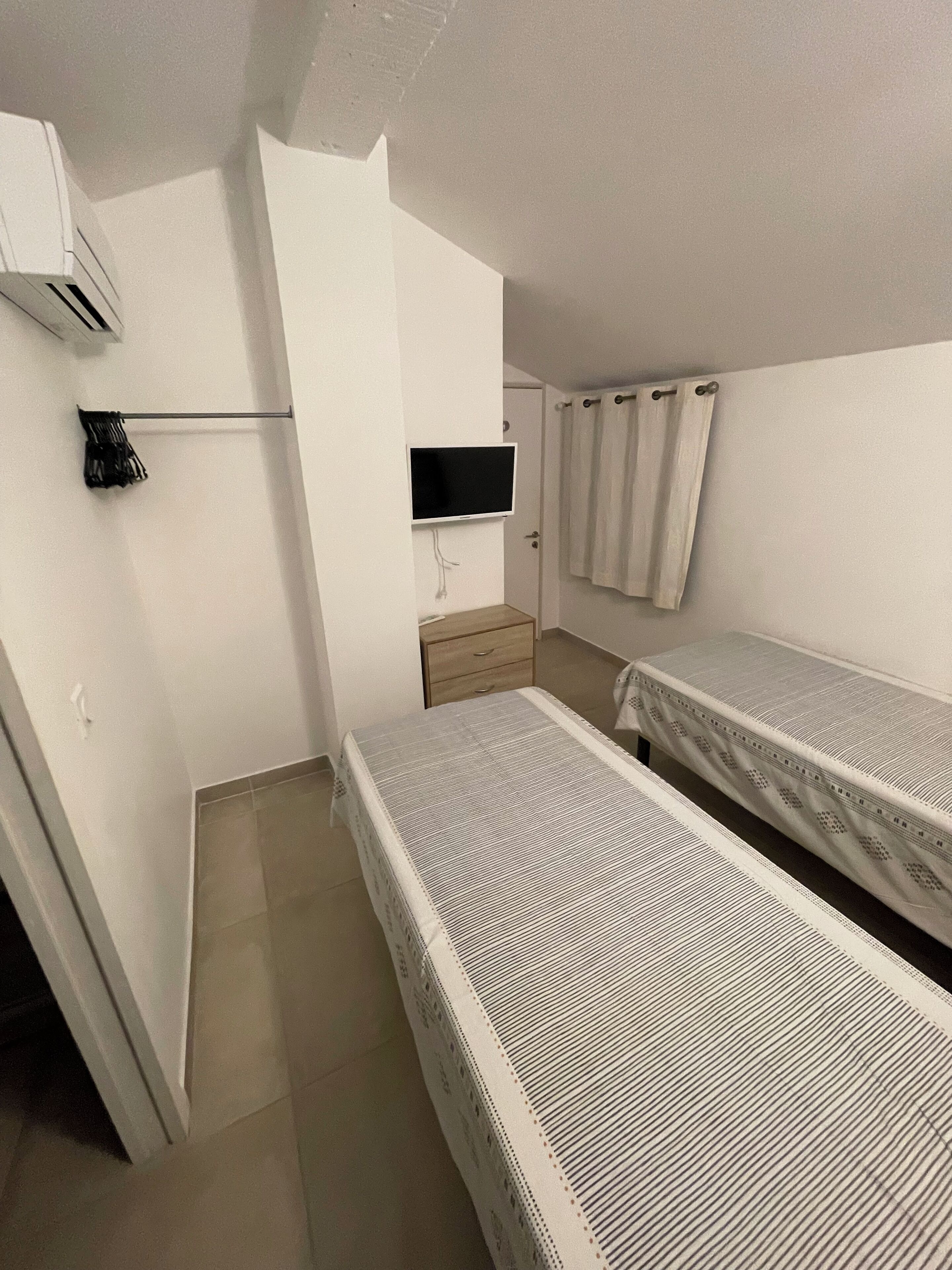 Une petite chambre propre, avec deux lits simples, un téléviseur fixé au mur, une petite commode en bois et un rideau à la fenêtre.