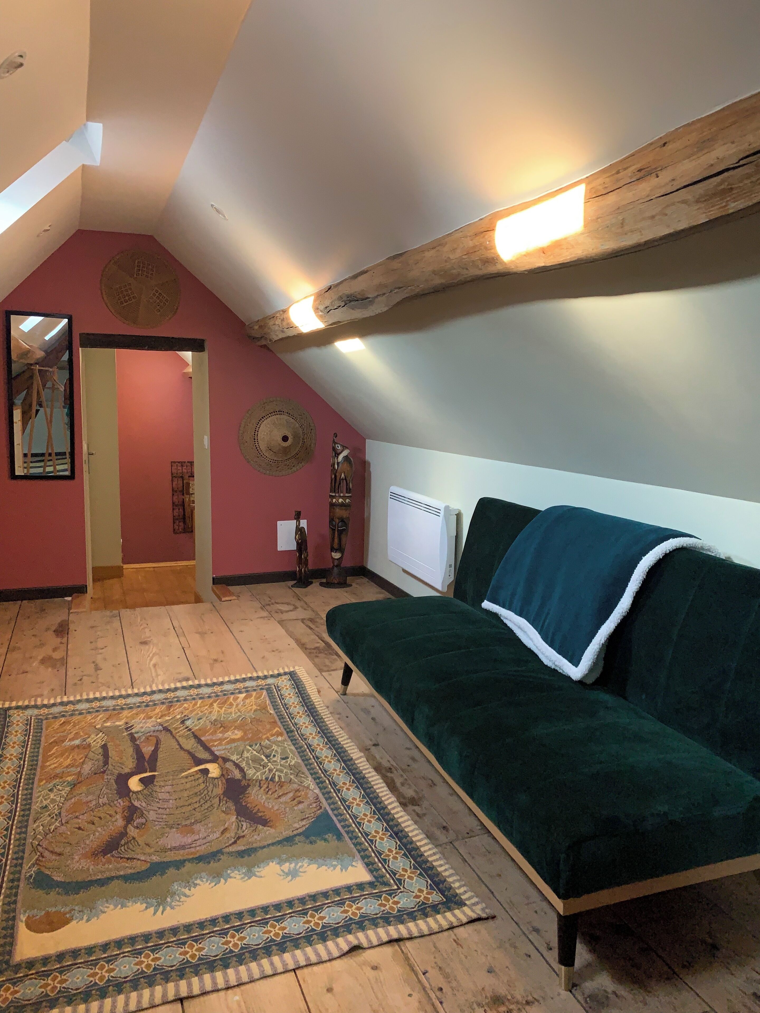 Une pièce mansardée chaleureuse, avec un canapé vert, un tapis à motifs, des poutres en bois et un miroir.