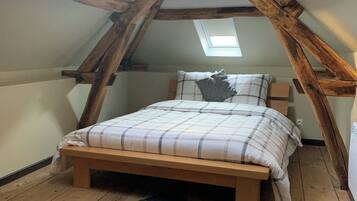 Une chambre à coucher aménagée dans un espace mansardé, avec un plafond en pente, des poutres en bois, un lit recouvert d’une couverture à carreaux et un tapis à motifs.