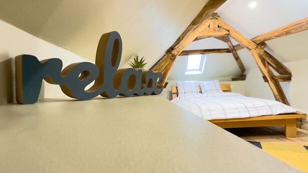 A cozy attic bedroom with a sloped ceiling, a bed with a white and yellow bedspread, and a large 'relax' sign. - Cocoon House – Private Garden & Exposed Beams – Peaceful Retreat 1h from Paris (Villeblevin)
