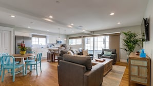 A spacious living area with a sofa, dining table, and chairs, a kitchen in the background, and large windows. - Seaglass Shores (San Diego)