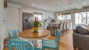 A modern kitchen with white cabinets, a stainless steel refrigerator, and a dining area with a round wooden table and blue chairs. - Seaglass Shores (San Diego)
