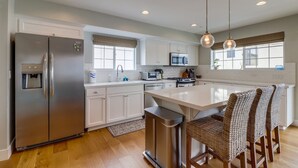 A modern kitchen with stainless steel appliances, white cabinetry, and a central island with wicker chairs. - Seaglass Shores (San Diego)