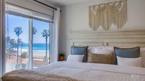 A bedroom with a large window overlooking a beach, a bed with multiple pillows, and a macrame wall hanging. - See the Sea 52 (San Diego)