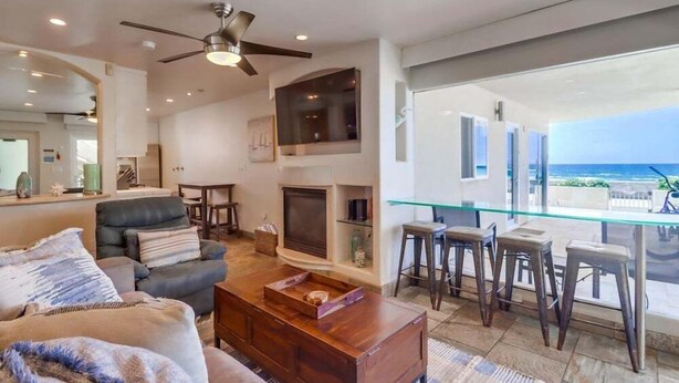 A spacious living room with a fireplace, a ceiling fan, and a glass dining area overlooking the ocean. - Surf Rider D (San Diego)