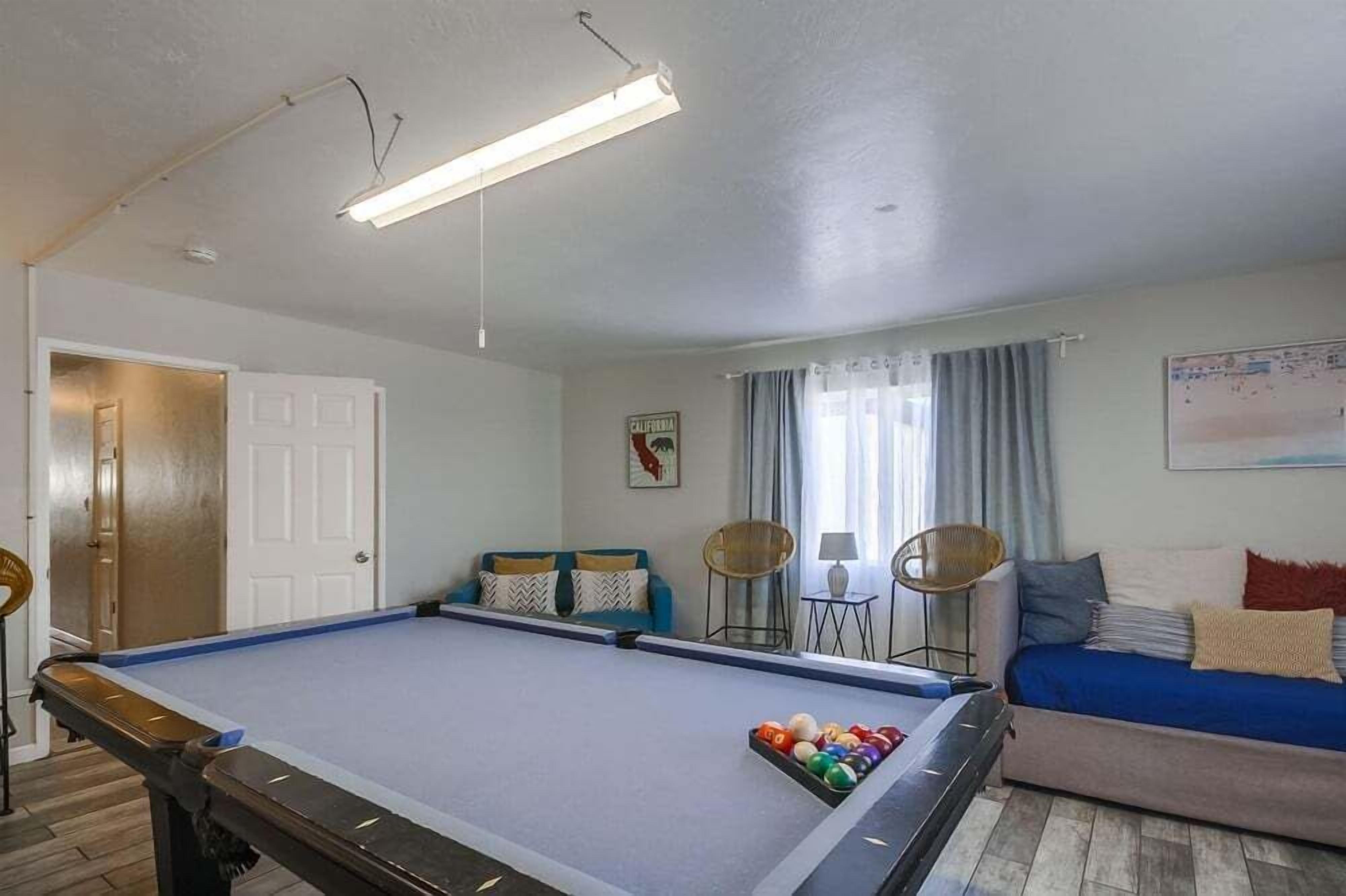 Ein Billardtisch mit Kugeln, eine Couch und zwei Sessel in einem Raum mit Tür und einem fenstervertäuschten Fenster.
