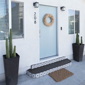 Une maison avec une porte bleu clair, le numéro 208, une guirlande, deux cactus en pot et une bordure de carreaux décoratifs.
