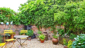 Un jardin avec un mur en briques, une chaise jaune, une table et diverses plantes en pot.