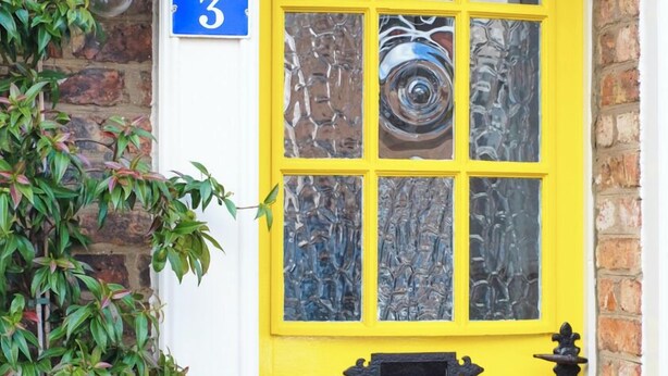 Une porte jaune vif avec un panneau en verre, une plaque de numéro de maison bleue et un pot de fleurs contenant des fleurs jaunes.