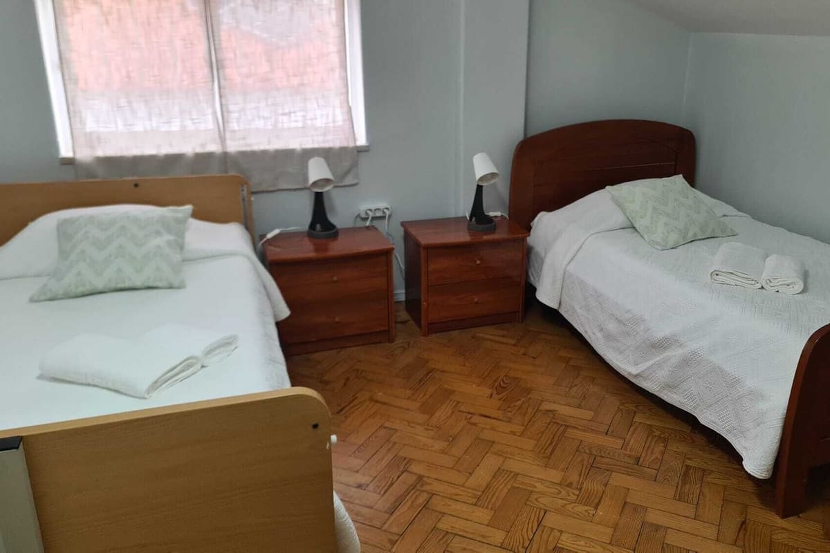 Una habitación pequeña y ordenada con dos camas de madera, una mesita de noche con lámpara y una ventana con cortinas de gasa.
