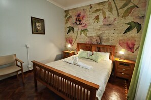 A neatly arranged bedroom with a wooden bed, a bench, a nightstand with a lamp, and a floral wallpaper. - Jardins de São Luís (Setubal)