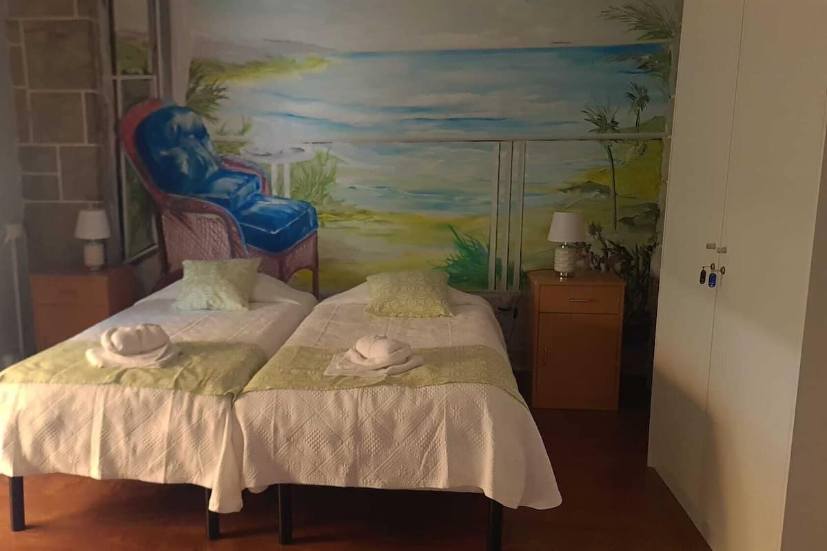 Habitación con dos camas, una silla, una mesita de noche y un mural con una playa.
