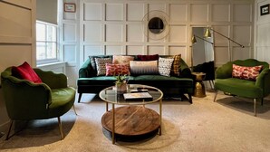 Een woonkamer met een groene bank, twee groene fauteuils, een ronde salontafel en een grote spiegel.