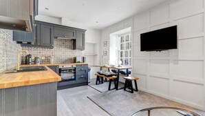 Een moderne keuken met grijze kastjes, een houten aanrechtblad en een ingebouwde oven. Er is een zithoek met een tafel en stoelen, en een televisie die aan de muur hangt.