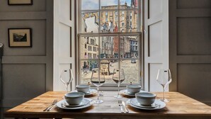 Mesa de jantar posta com pratos, tigelas e copos de vinho, posicionada em frente a uma janela com vista para a cidade.