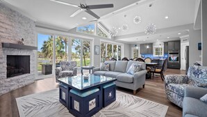 A spacious living room with a fireplace, a glass-top coffee table, and a large window overlooking a garden. - Brand New, Golf Cart Included - 546 Remora Dr. (Fripp Island)