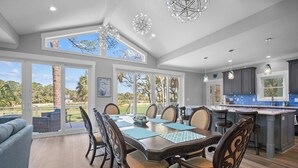 A spacious dining area with a large table and chairs, a kitchen with dark cabinets and blue backsplash, and large windows offering a view of the outdoors. - Brand New, Golf Cart Included - 546 Remora Dr. (Fripp Island)