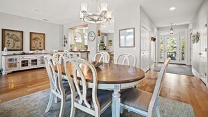 A spacious dining area with a wooden table and white chairs, a chandelier, and a view of the kitchen and dining room in the background. - Lakefront, Golf Cart Included, Pet Friendly - 259 Deer Run (Fripp Island)