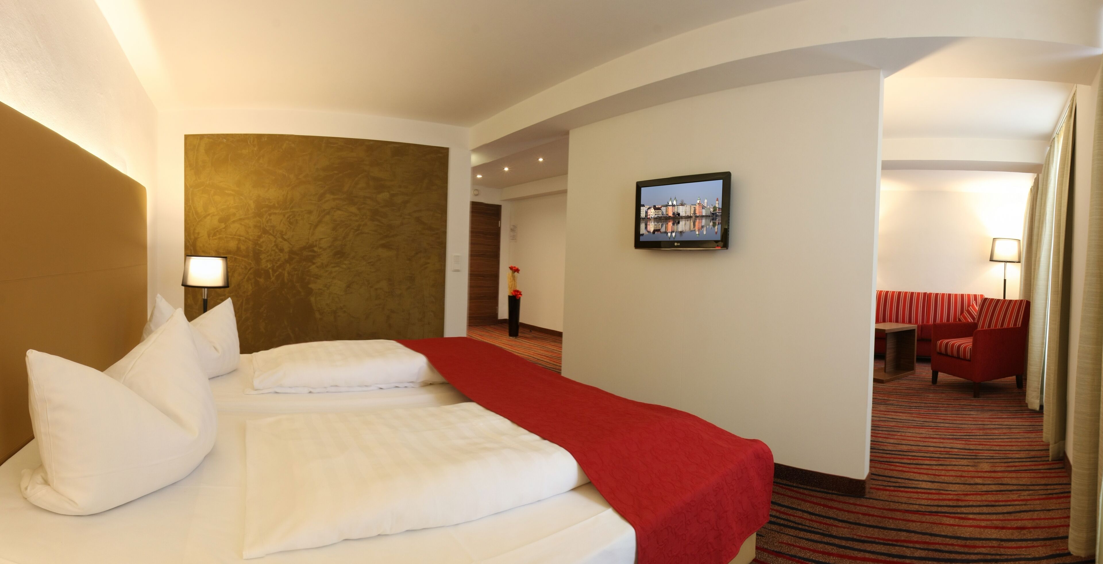 Ein Hotelzimmer mit einem großen Bett, einer roten Tagesdecke, einem Flachbildfernseher an der Wand und einem rot gestreiften Sofa in der Ecke.