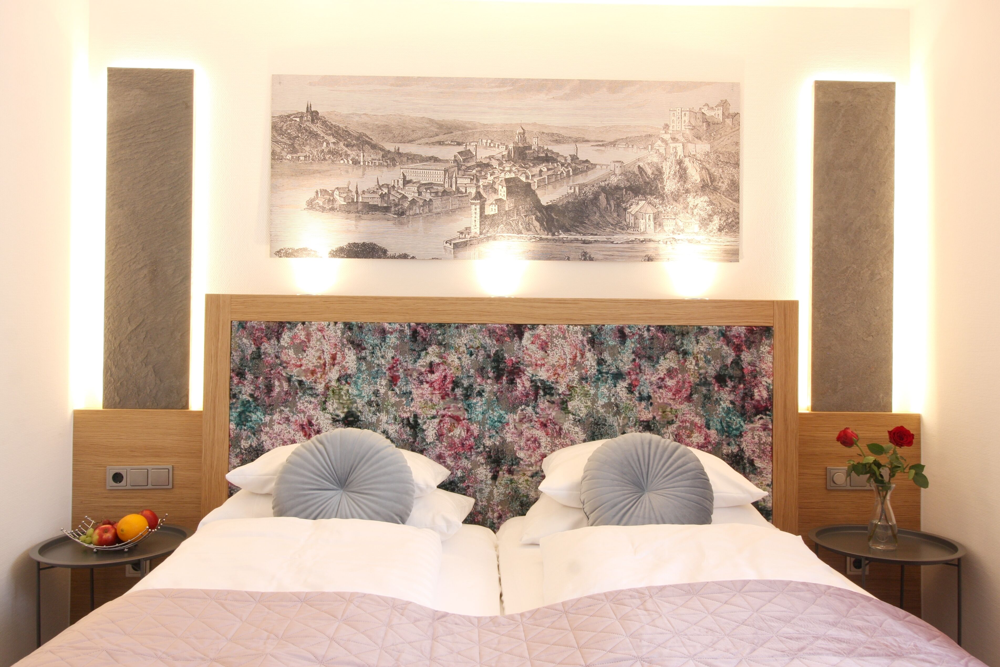 Ein Hotelzimmer mit einem großen Bett, einem blumigen Kopfstück, einem Stadtbild über dem Bett und einem kleinen Tisch mit Blumen.'