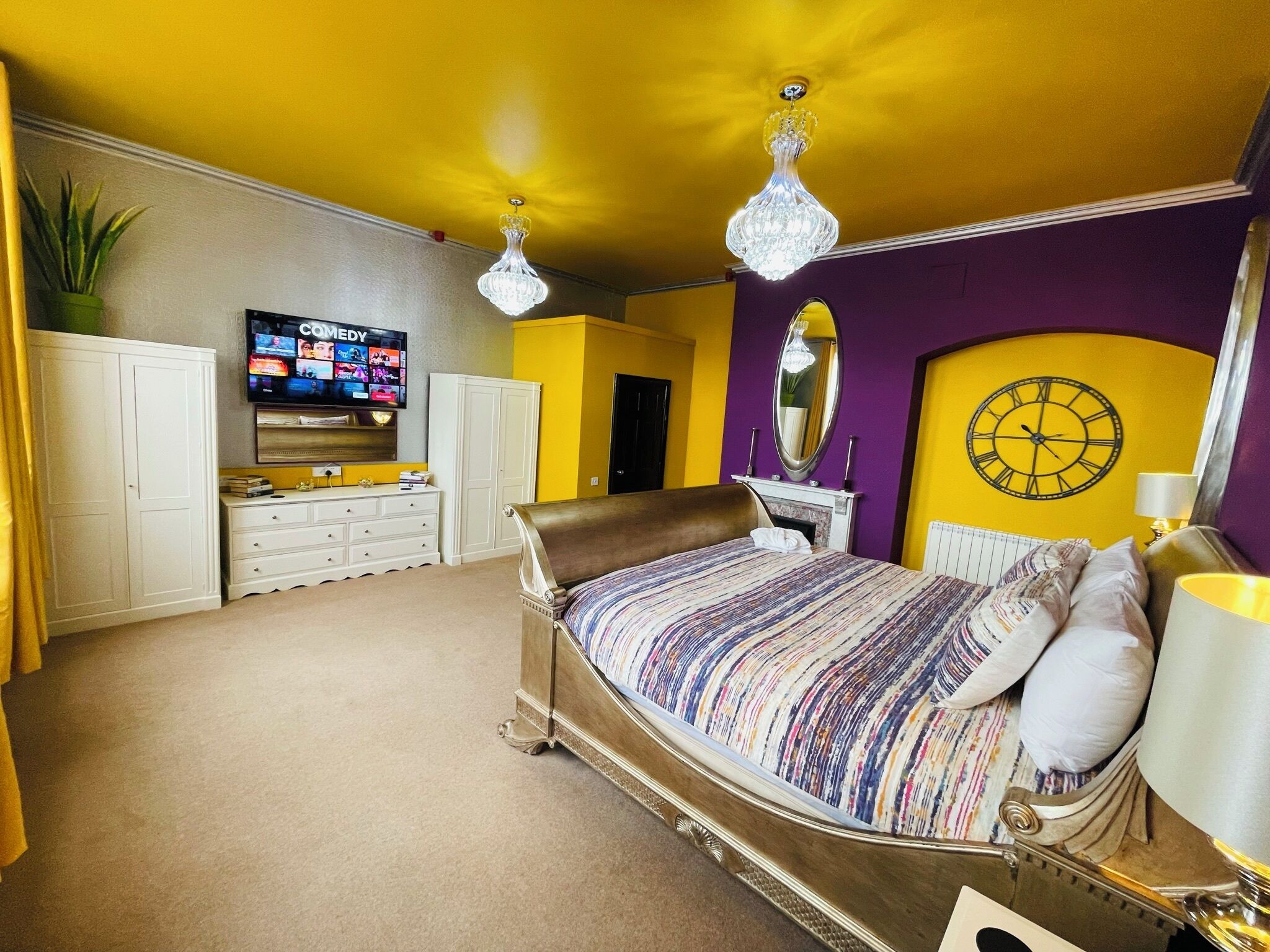 A bedroom with a bed, dresser, vanity, mirror, and television.