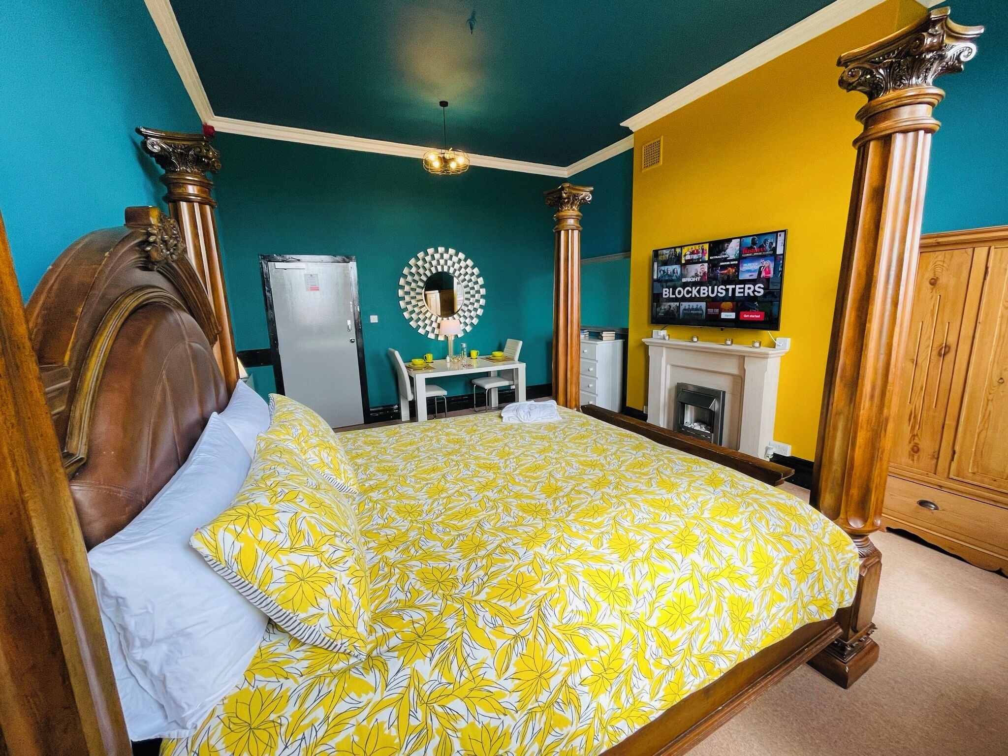 A bedroom with a four-poster bed, a small table with chairs, a fireplace, and a wall-mounted TV displaying 'BLOCKBUSTERS'.