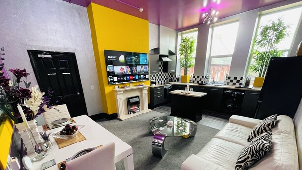 A modern living room with a white sofa, a glass coffee table, and a dining area with a white table and chairs. There is a fireplace, a kitchen area with a checkered backsplash, and a large window with a view of greenery. - PHOENIX Apartments (Goole)