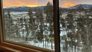 Une vue depuis une fenêtre donnant sur un paysage enneigé, avec des arbres et des montagnes à l’horizon.