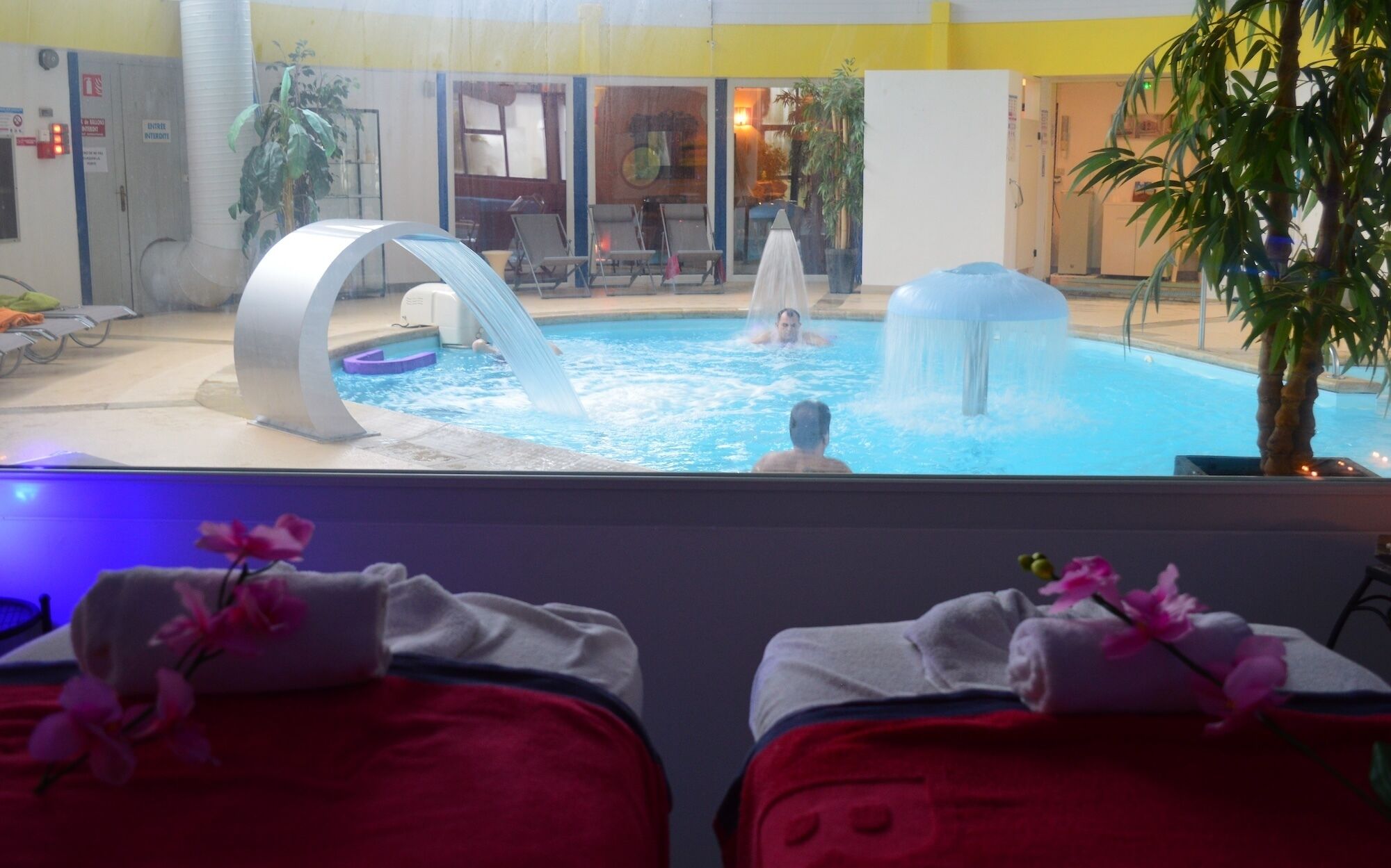 Un espace piscine agrémenté d’un élément décoratif aquatique, de chaises longues et offrant une vue sur la piscine depuis une chambre dotée d’un lit et d’une plante.
