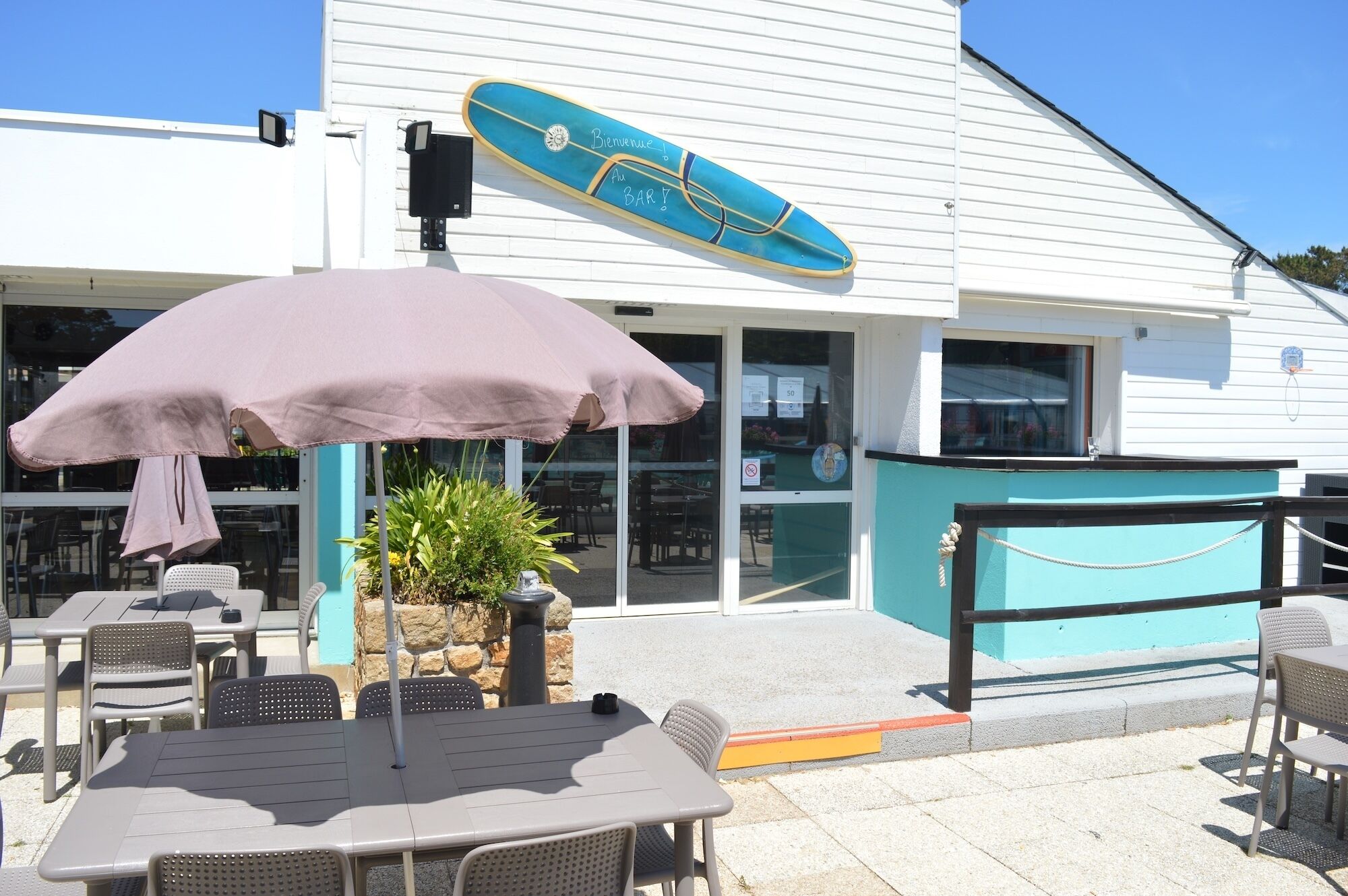 Een buitenzitplaats met tafels en stoelen, een potplant en een blauw surfboard met tekst.