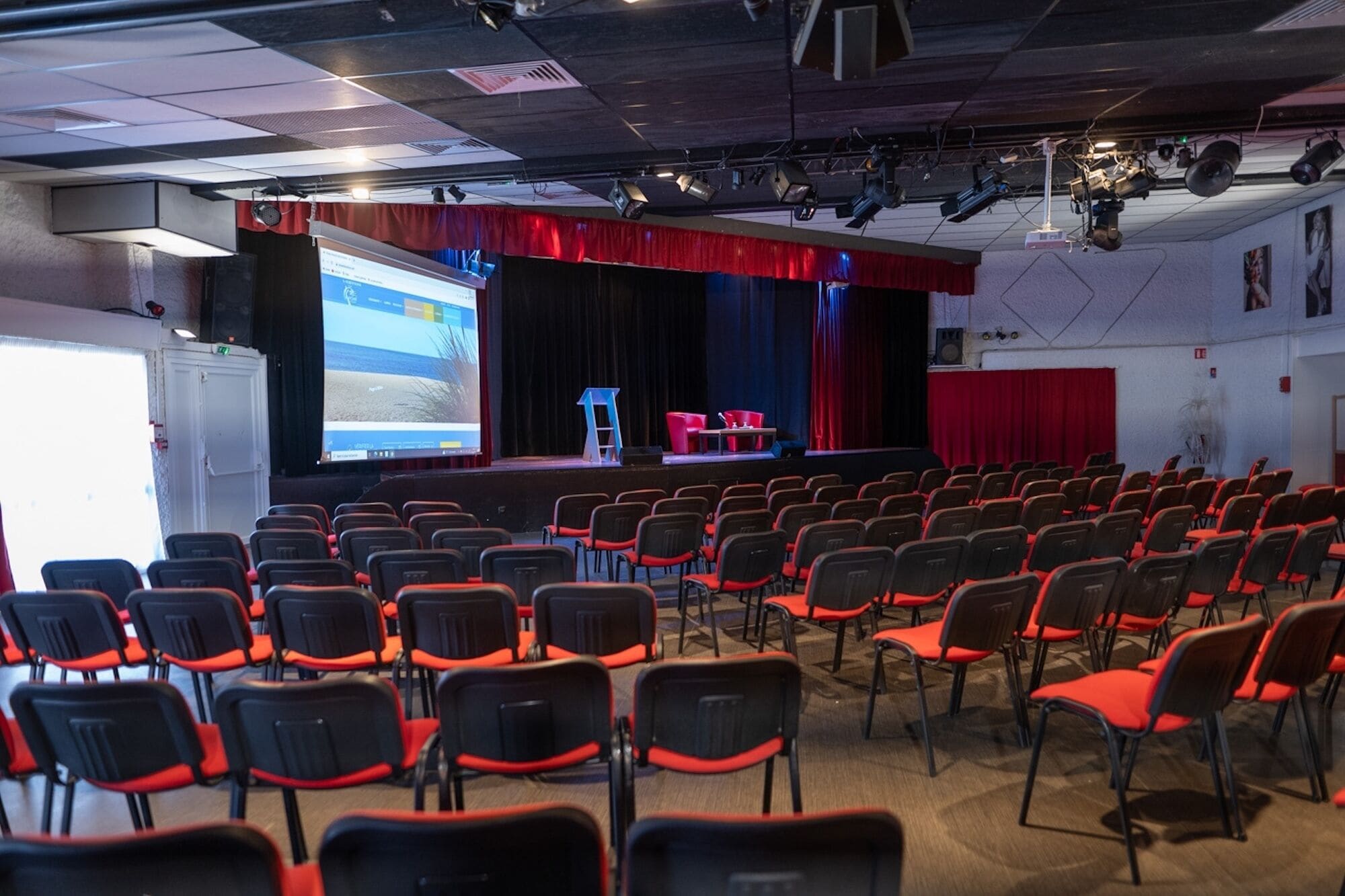 Un auditorio con filas de sillas, un escenario con cortina roja y una pantalla de proyección.
