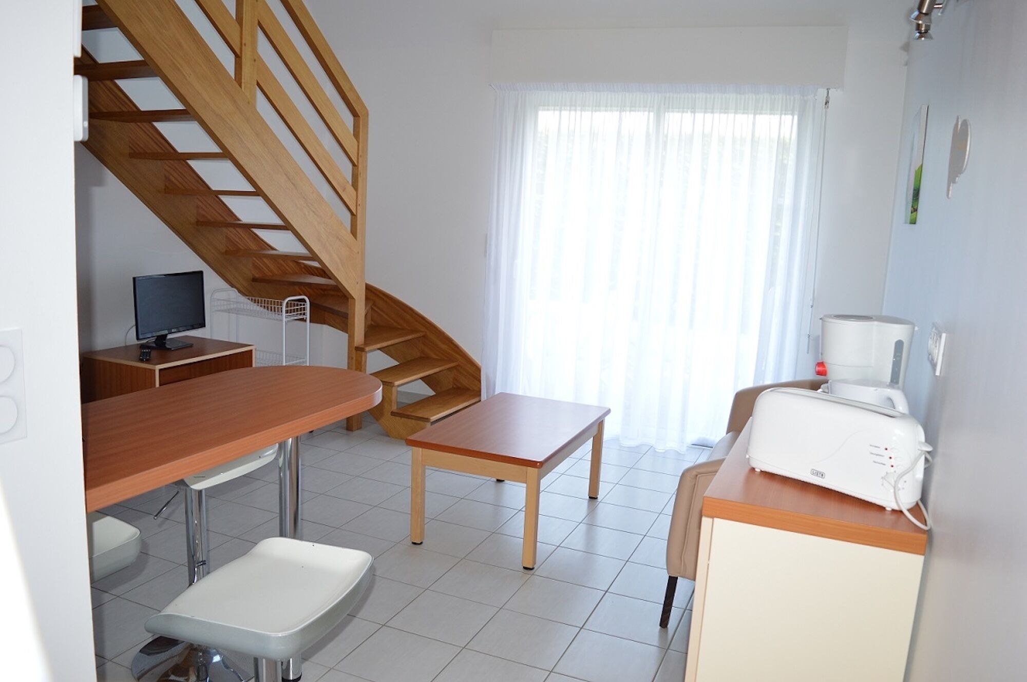 Ein Zimmer mit einer Holztreppe, einem Schreibtisch mit Computer, einem kleinen Tisch, einem Stuhl, einem Bett und einem Fenster mit Vorhängen.