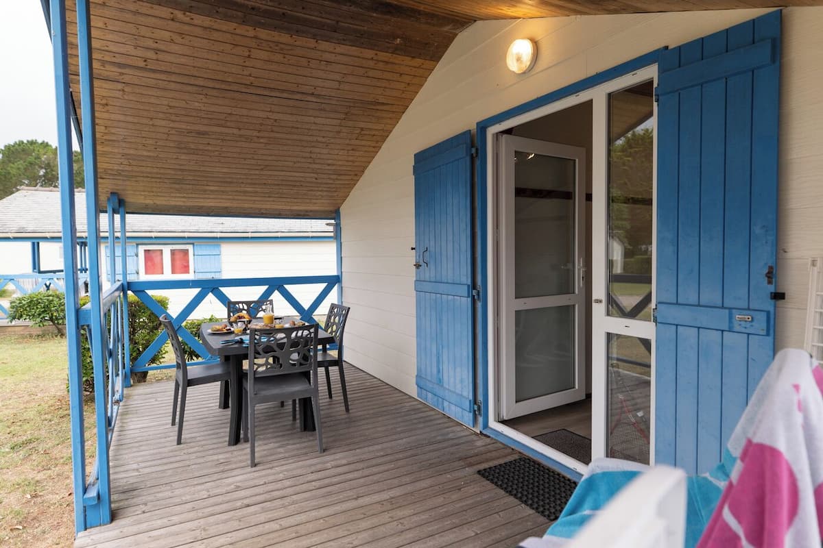 Een overdekt buitengebied met een tafel en stoelen, een houten terras en een schuifdeur die naar een gebouw leidt.
