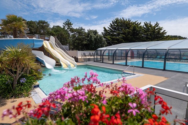 Una piscina con tobogán, zona cubierta y vegetación circundante.