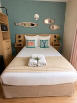 A neatly made bed with striped bedding, two pillows with marine-themed designs, a bedside table with a lamp, and a wall-mounted mirror.