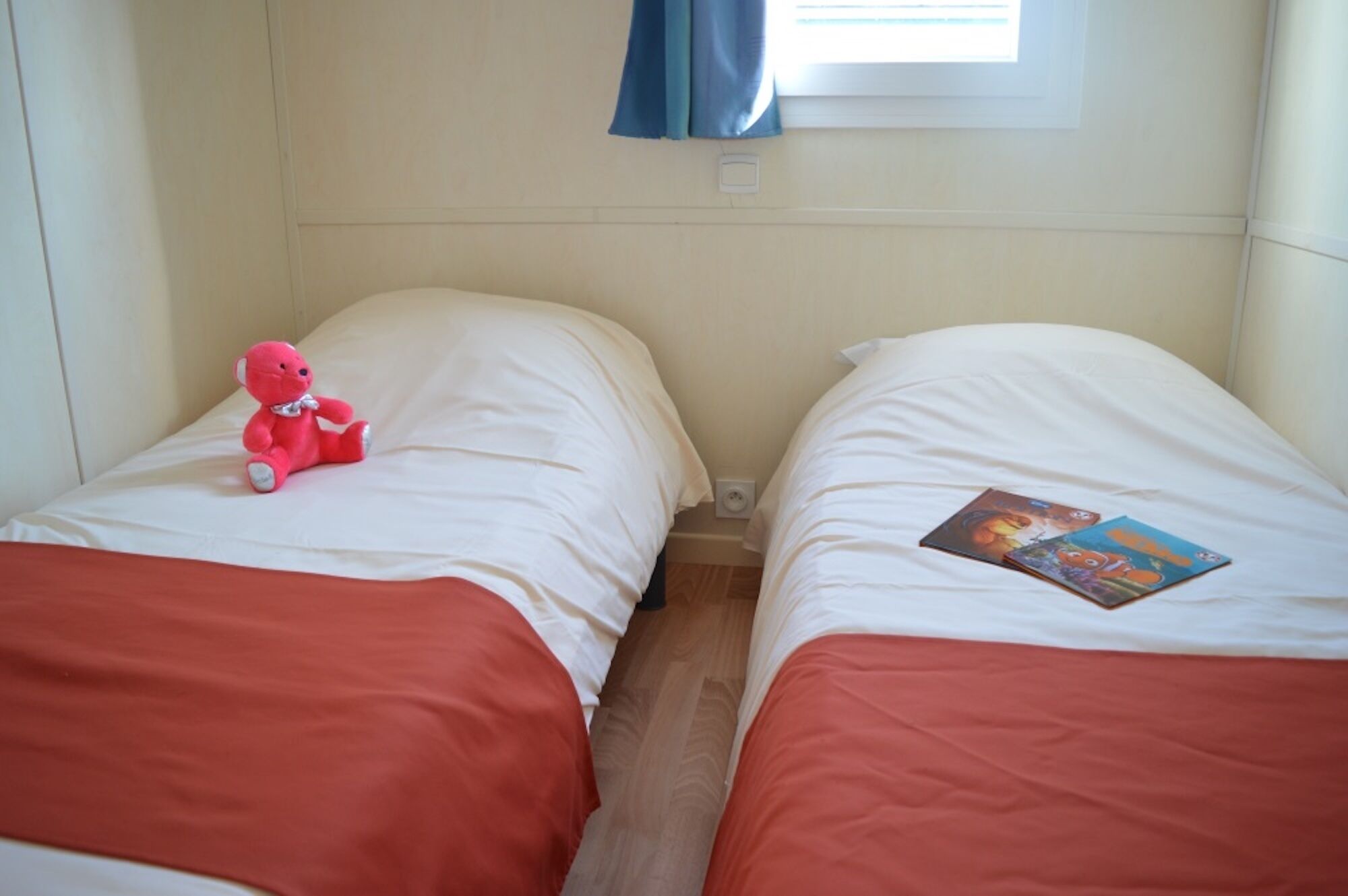 Deux lits simples avec une literie blanche et des têtes de lit rouges, un ours en peluche rose sur le lit de gauche et un livre sur le lit de droite.
