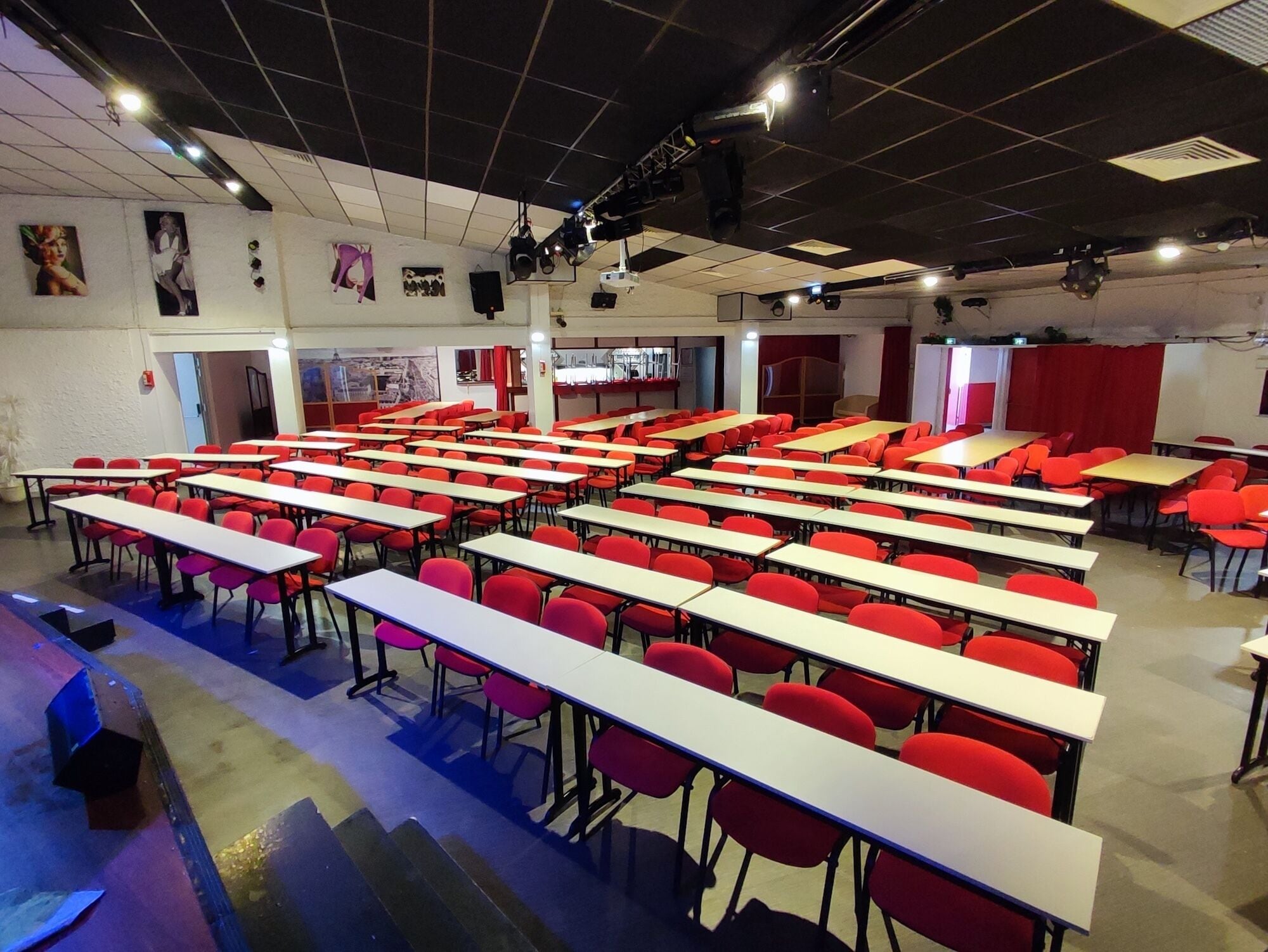 Ein Klassenzimmer mit Reihen von Schreibtischen und Stühlen, eine Bühne mit Beleuchtungstechnik und eine Tür zu einem anderen Raum.