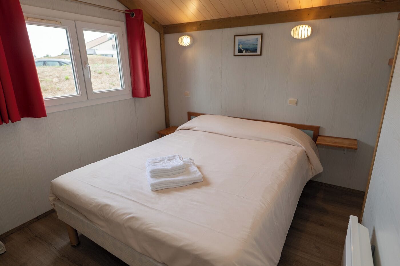 Un dormitorio con cama, una ventana con cortinas, una mesita de noche y una lámpara de pared.