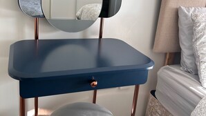 A room with a blue vanity, a round mirror, a small stool, and a bed with patterned bedding.