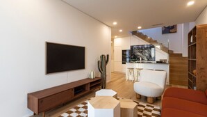 Un salon moderne doté d’un téléviseur à écran plat, d’un meuble de rangement en bois, d’un cactus, d’un fauteuil blanc et d’un escalier menant à la cuisine.
