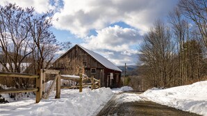 A rustic barn with a snow-covered roof, surrounded by bare trees and a wooden fence.