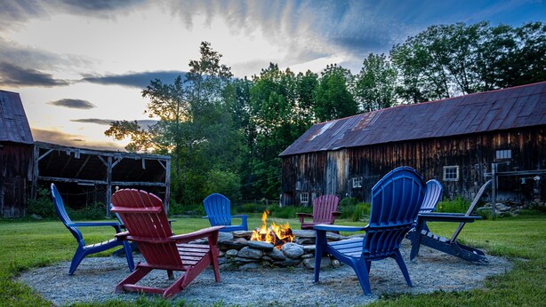 A rustic outdoor seating area with Adirondack chairs around a campfire, set against a backdrop of barns and trees.