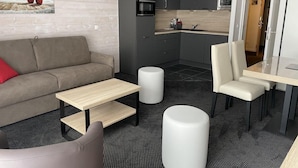 Un salon moderne avec un canapé, une table basse et un pouf, une cuisine avec des meubles gris et un four à micro-ondes, et un coin repas avec une table et des chaises.