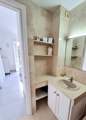 A bathroom with beige marble walls, a white sink, and shelves with toiletries.