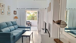 Una sala de estar moderna con un sofá azul, una mesita y un televisor. Hay cuadros en la pared y una ventana con cortinas.