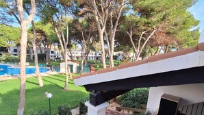 Un balcón con mesa y sillas, con vista a una piscina y un jardín bien cuidado con árboles y césped.