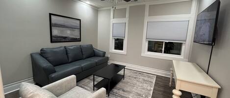 Una sala de estar con un sofá de cuero oscuro, un sillón de color claro, una mesa de centro, un televisor sobre un mueble bajo y un ventilador de techo.