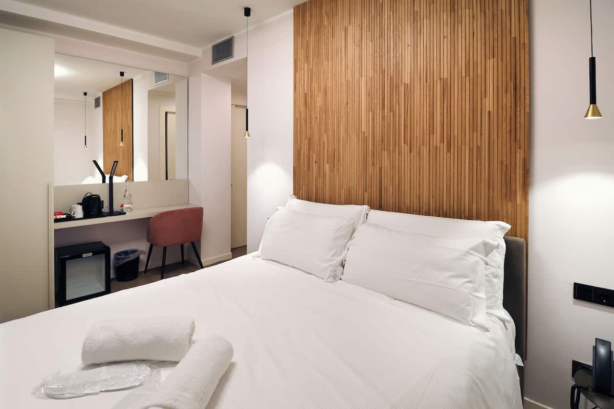 A modern hotel room with a large bed, a wooden headboard, a desk with a chair, a mirror, and a small TV.