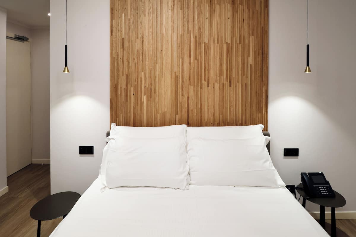 A hotel room with a wooden headboard, white bedding, a black bedside table with a phone, and pendant lighting.